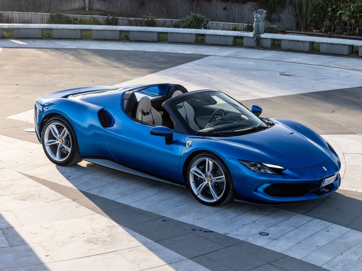 A bright blue Ferrari convertible with tan interior facing a corner, parked on a light-colored paved space with shadows.