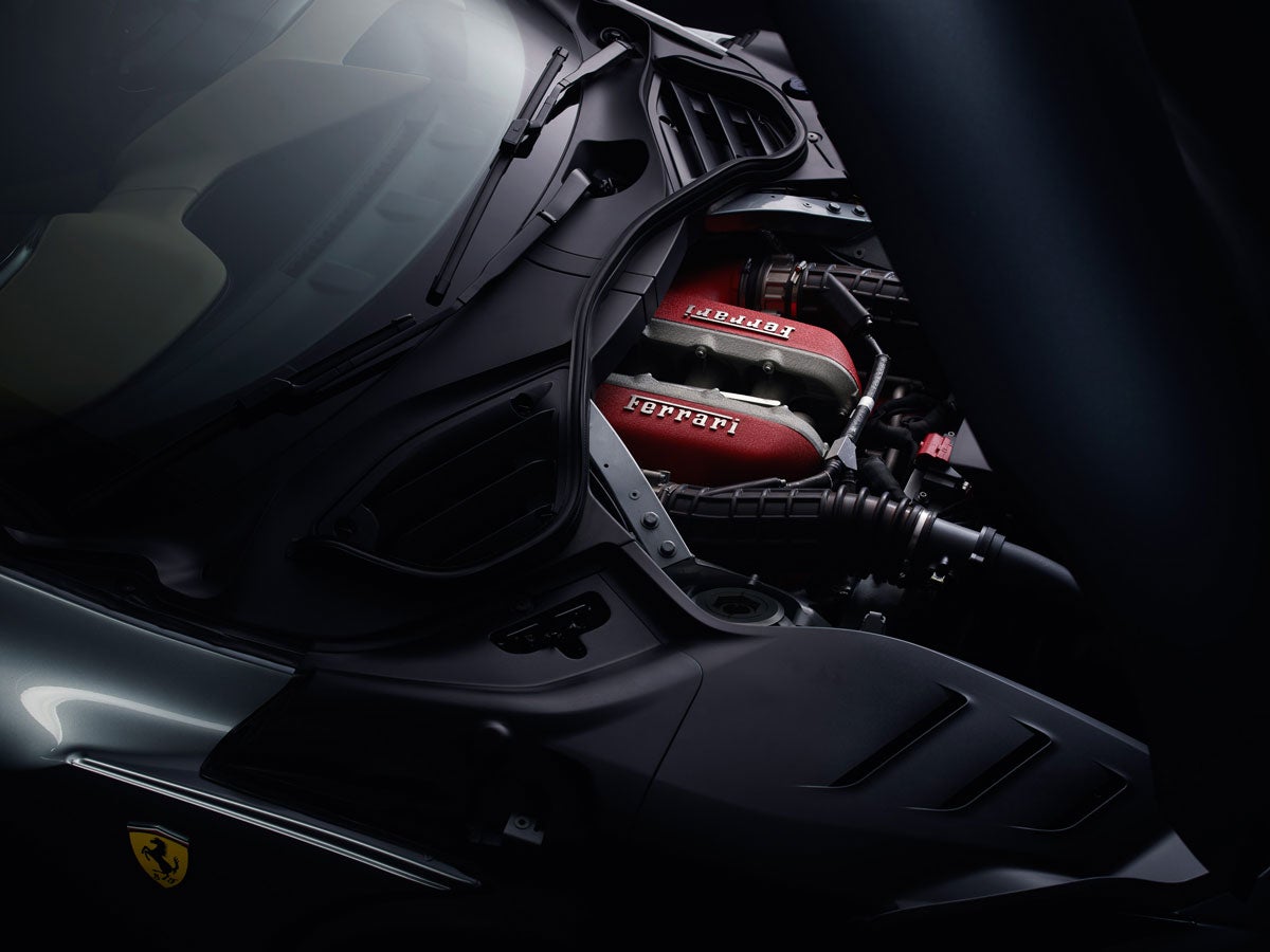 The red engine of a Ferrari is revealed under the partially opened dark gray hood of the car.